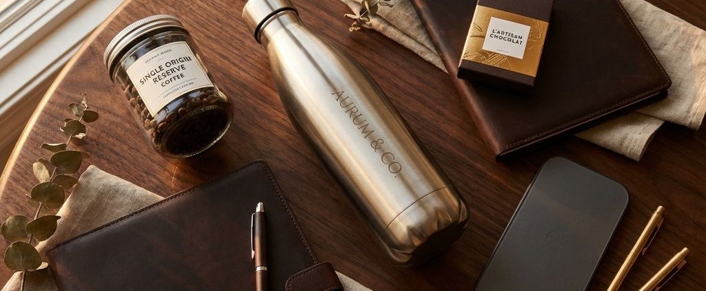 Luxe bedrijfscadeaus op donker hout: roestvrijstalen waterfles, leren portfolio, koffie en chocolade in gouden licht