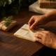 Handen die elegante goudfolie branding aanbrengen op witte eco-vriendelijke voedselverpakking op houten bureau met dennentakjes