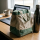 Versleten canvas lunchtas met zichtbare plooien naast waterles op modern kantoorbureau tussen laptop en koffiemok