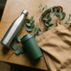 Duurzame relatiegeschenken: roestvrijstalen drinkfles, matte thermobeker en branded tote bag op eiken bureau met eucalyptustakjes.