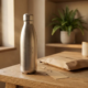 Gegraveerde roestvrijstalen drinkfles op eikenhouten bureau met kraftpapier en cadeaulabel, zachte kantoorsfeer met groene plant op achtergrond.