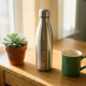 Gepersonaliseerde roestvrijstalen herbruikbare drinkfles op houten bureau naast succulent en keramische koffiemok, warm ochtendlicht.