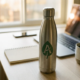Gepersonaliseerde roestvrijstalen herbruikbare waterfles op modern bureau met laptop en koffie op de achtergrond.