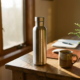 Roestvrij stalen herbruikbare waterfles op een eiken bureau naast een keramische koffiemok en groen plantje, warm middaglicht.