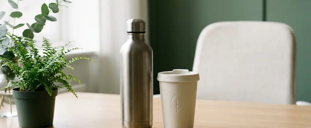 Roestvrijstalen herbruikbare waterfles met matte afwerking op minimalistisch bureau naast keramieken reismok, omringd door groen.