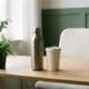Roestvrijstalen herbruikbare waterfles met matte afwerking op minimalistisch bureau naast keramieken reismok, omringd door groen.