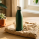 Roestvrij stalen herbruikbare waterfles op linnen cadeaupapier naast een kleine vetplant, warm ochtendlicht op wit eikenhouten bureau.