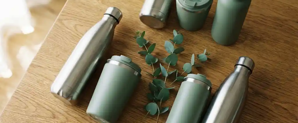 Herbruikbare roestvrijstalen waterflessen en geïsoleerde koffiebeker in saliegroen en zilver op eiken ondergrond met eucalyptustakje.