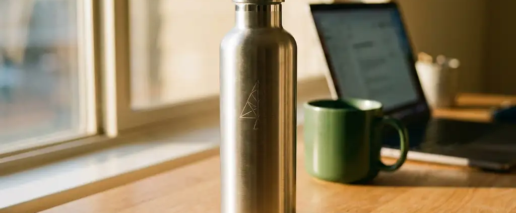 Roestvrijstalen herbruikbare waterfles op een modern bureau, met een laptop en koffiemok op de achtergrond in warm goudkleurig licht.