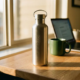 Roestvrijstalen herbruikbare waterfles op een modern bureau, met een laptop en koffiemok op de achtergrond in warm goudkleurig licht.