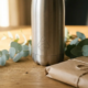 Gepersonaliseerde roestvrijstalen herbruikbare waterfles met gegraveerd logo op eikenhouten bureau, omringd door eucalyptustakjes en kraftpapier.