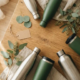 Herbruikbare roestvrijstalen waterflessen en thermokoffiebekers op eiken ondergrond met eucalyptustakjes en linnen stof.