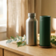 Roestvrijstalen herbruikbare waterfles en matte thermosbeker op licht eiken bureau met groene plantentakjes in warm daglicht.
